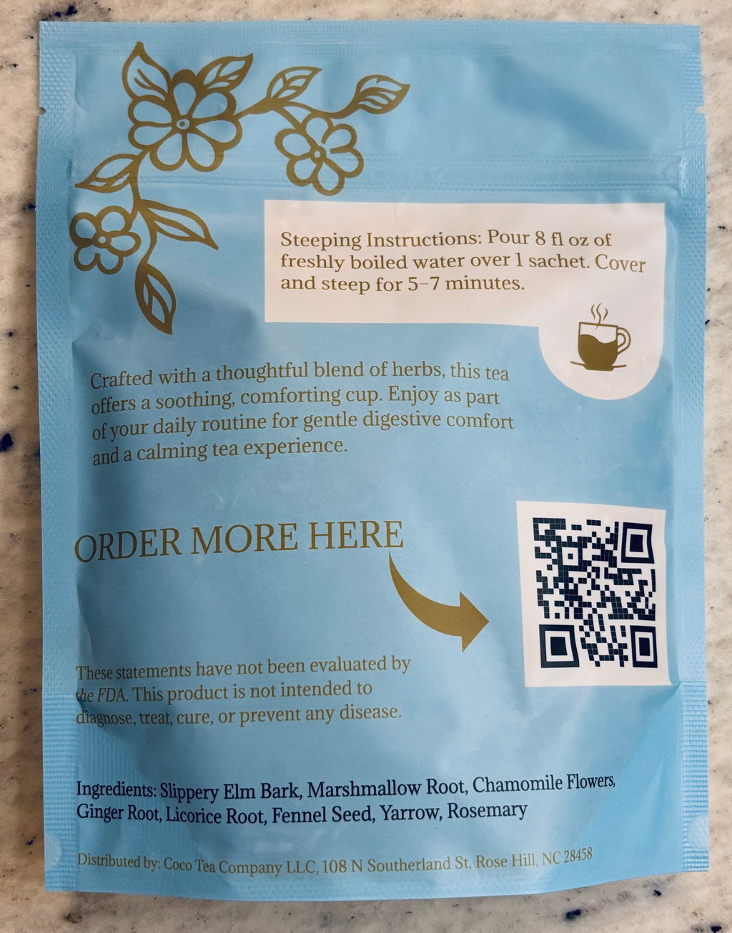 A light blue tea package with gold floral illustrations and black text, including steeping instructions and a QR code, photographed flat against a light neutral surface.
