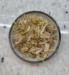 A top-down view of loose-leaf herbal tea, featuring dried yellow flowers and green leaves mixed with fibrous stems, presented in a small ceramic bowl on a speckled white marble surface.
