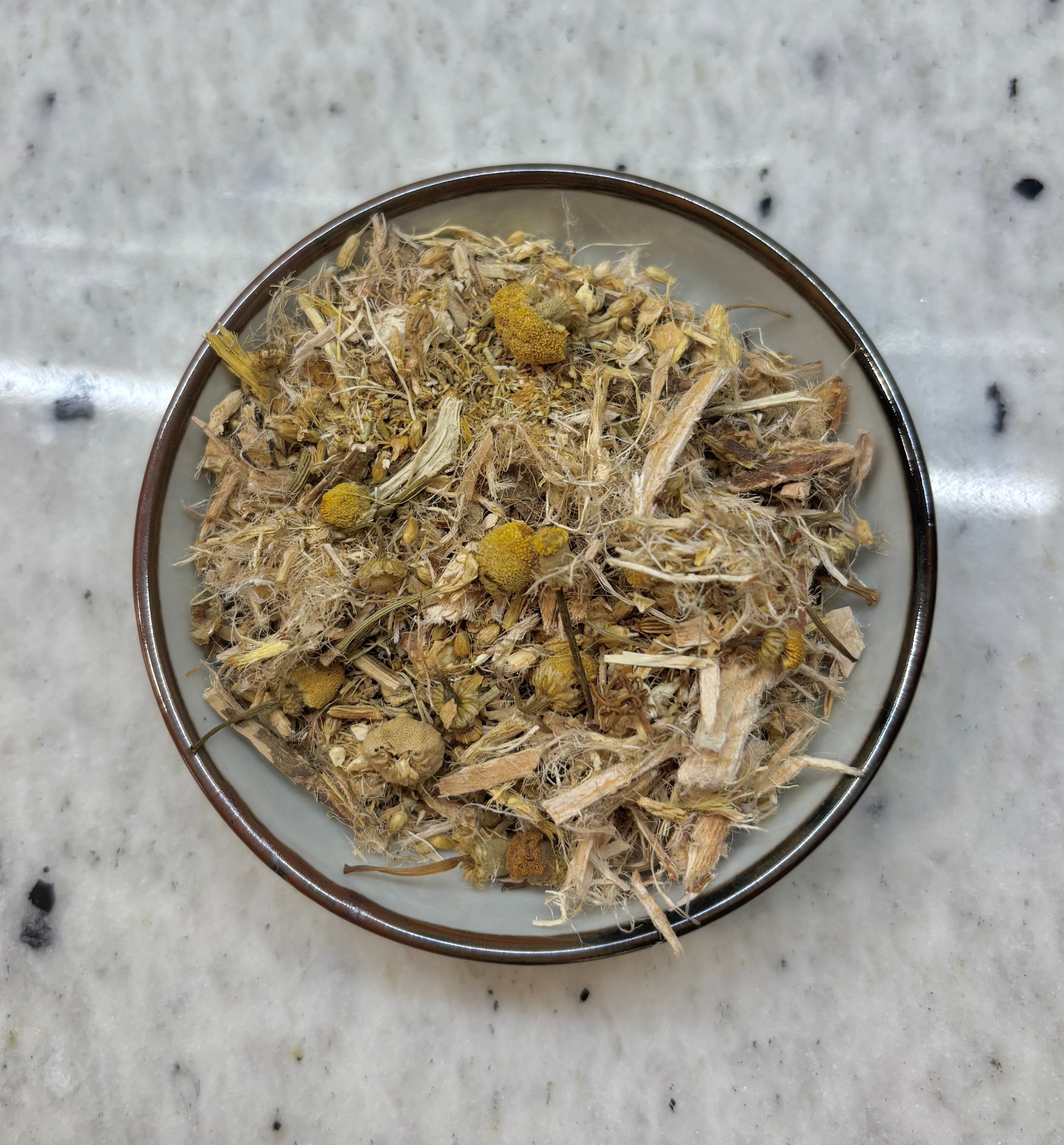 A top-down view of loose-leaf herbal tea, featuring dried yellow flowers and green leaves mixed with fibrous stems, presented in a small ceramic bowl on a speckled white marble surface.