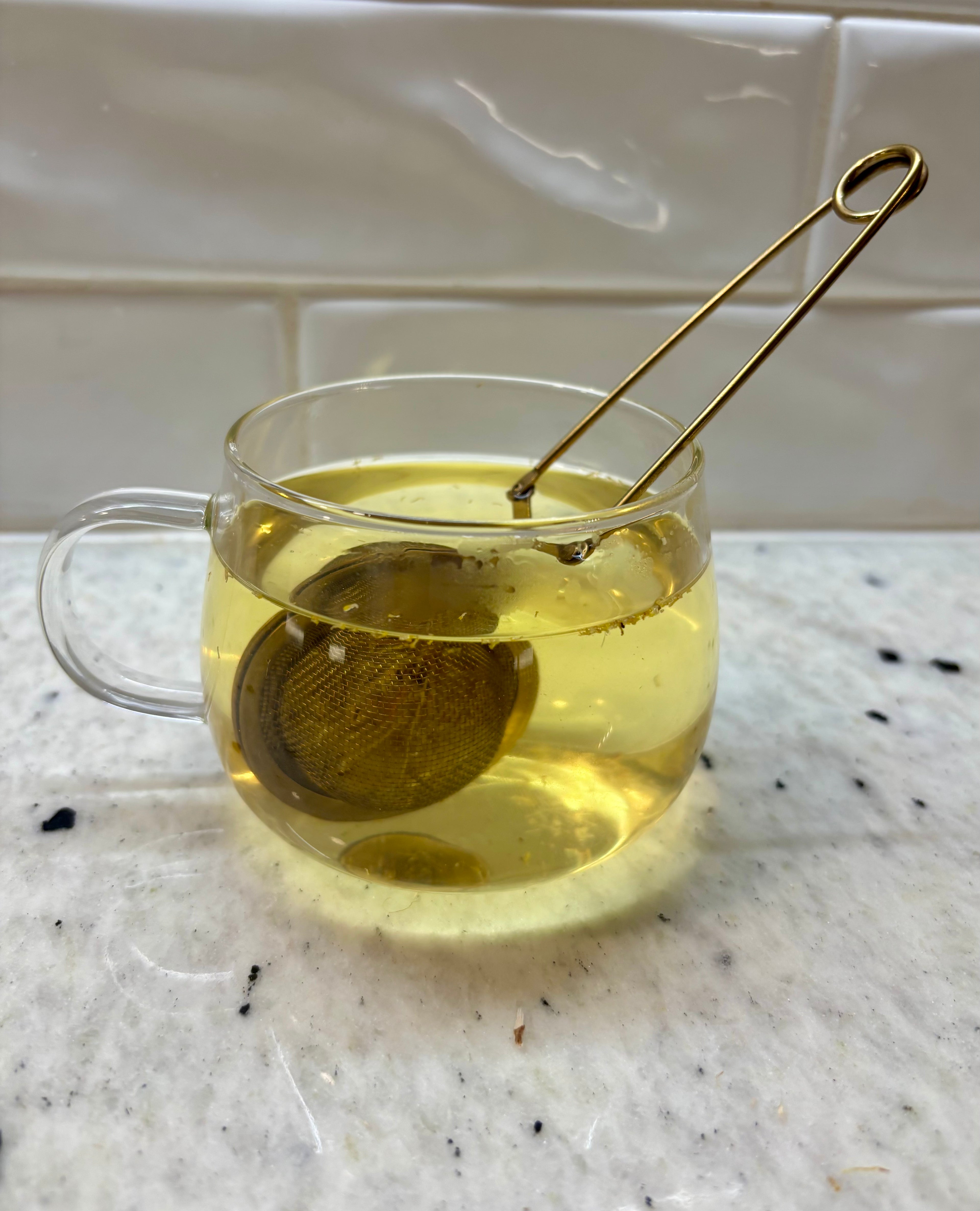 A clear glass teacup with a handle on a speckled white marble countertop, filled with pale yellow tea and containing a gold metal tea infuser. A white subway tile backsplash is visible in the background.