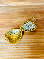A brass heart-shaped tea infuser with a gold-toned metal body, shown open on a wooden tabletop with loose tea visible inside.