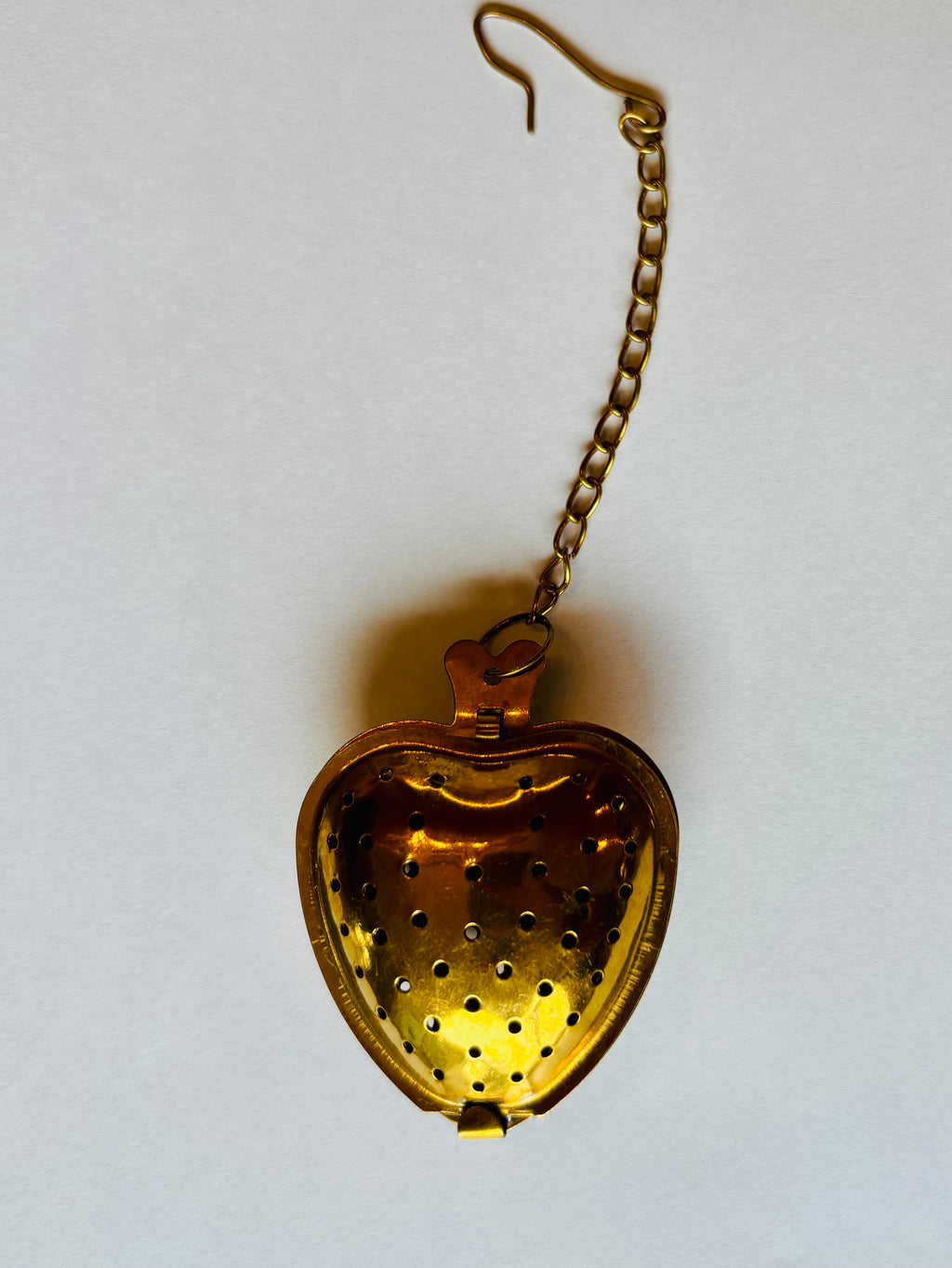 A vintage gold-toned metal heart-shaped tea infuser or strainer with a hanging chain, shown open against a plain off-white background.