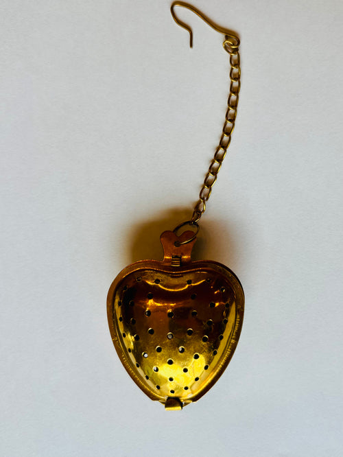 A vintage gold-toned metal heart-shaped tea infuser or strainer with a hanging chain, shown open against a plain off-white background.