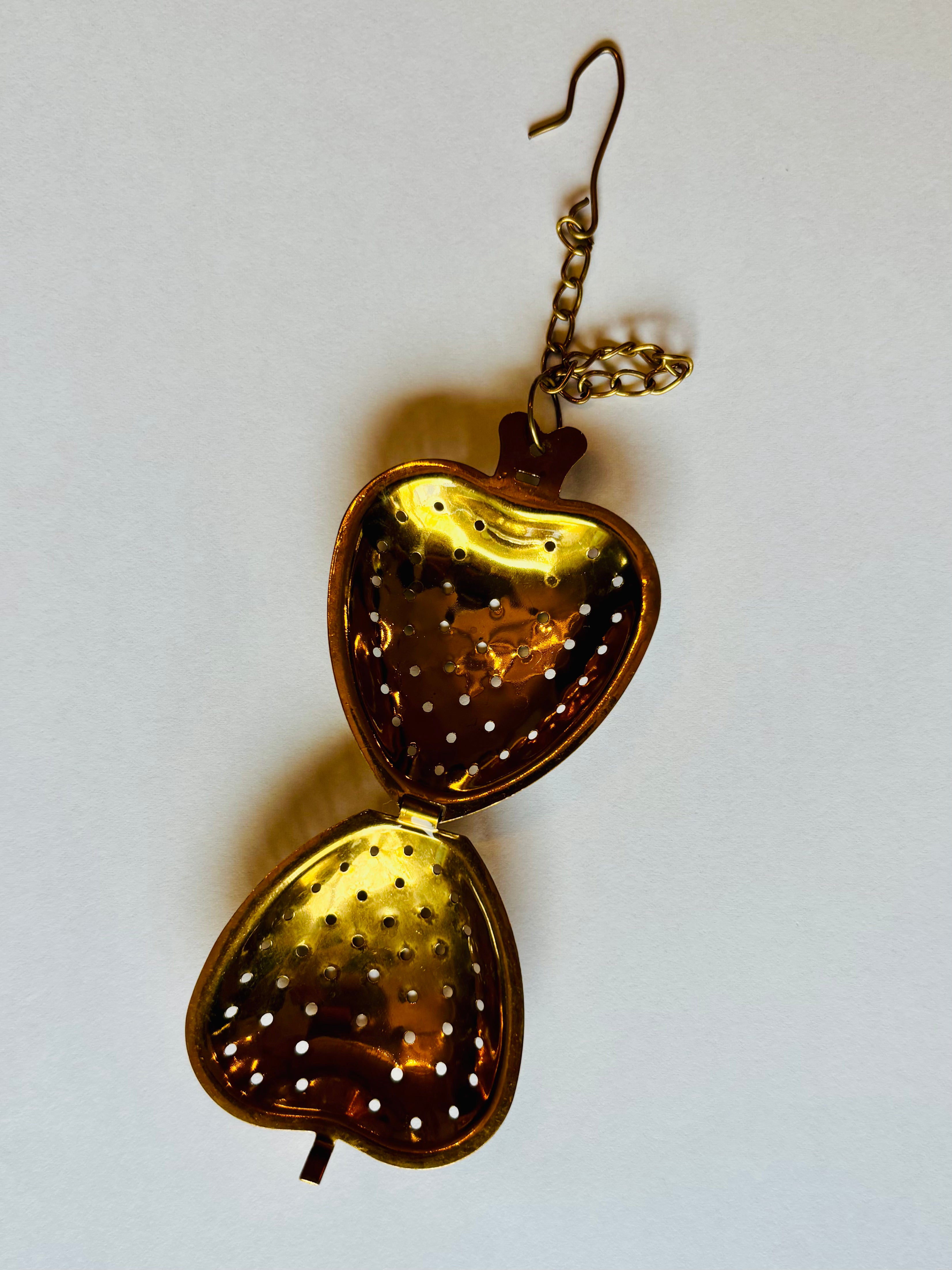 A vintage-style gold heart-shaped tea strainer with a hanging chain, shown open against a plain off-white background.